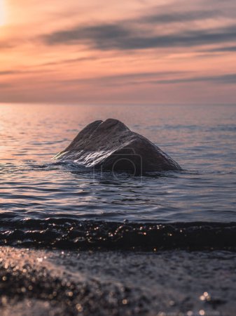 As the sun sets on Veczemju Klintis, a lone stone stands silently amidst the gentle waves, reflecting the colors of twilight in tranquil serenity.