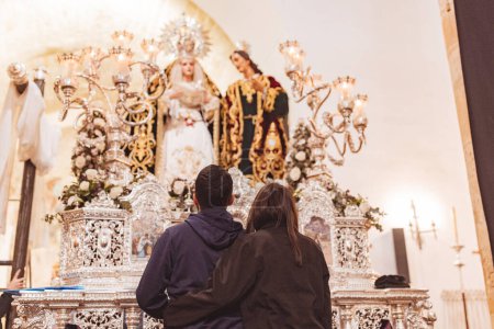 Téléchargez les photos : Couple avec le dos à l'autre et embrassant contemplant une statue de la Vierge Marie sur un trône de la Semaine Sainte. - en image libre de droit