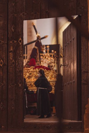 Téléchargez les photos : Porte entrouverte révèle le trône de la Semaine Sainte avec des pénitents à l'arrière de l'église - en image libre de droit