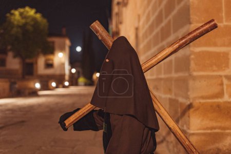 Téléchargez les photos : Nazaréen vêtu de tunique brune et visage couvert marchant et traînant une grande croix la nuit sous la lune à travers une vieille ville espagnole. - en image libre de droit