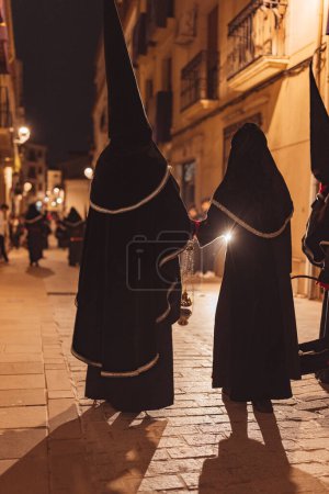Téléchargez les photos : Pénitents en procession la nuit avec le dos tourné en tunique à capuche noire tenant un encensoir - en image libre de droit