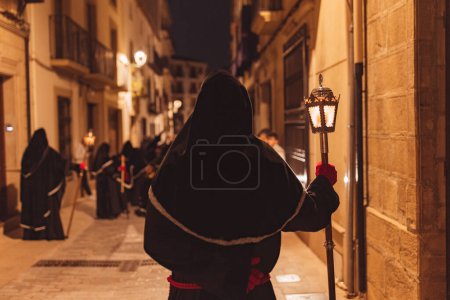 Téléchargez les photos : Pénitent en procession la nuit sur le dos avec une tunique noire et des gants rouges tenant une lanterne allumée. - en image libre de droit