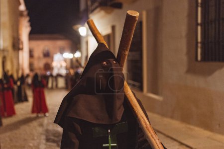 Téléchargez les photos : Pénitent au premier plan marchant avec une croix de bois sur les épaules dans une rue de pierre la nuit. - en image libre de droit