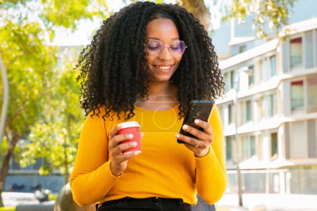 Téléchargez les photos : Adolescent heureux, boire du café envoyer des messages à l'aide d'un téléphone portable à l'extérieur. Communication - en image libre de droit