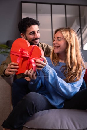Foto de Hermosa pareja joven en casa disfrutando del tiempo juntos mientras celebran el Día de San Valentín con una caja de regalo en forma de corazón en la mano. - Imagen libre de derechos