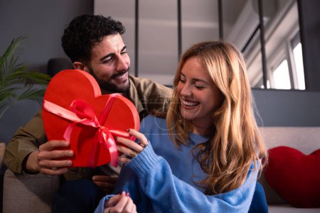 Téléchargez les photos : Beau jeune couple souriant à la maison profitant du temps ensemble tout en célébrant la Saint-Valentin avec une boîte-cadeau en forme de coeur à la main. - en image libre de droit