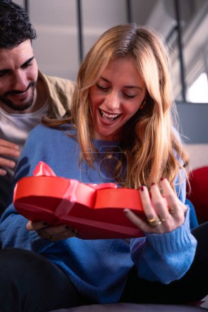 Foto de Hermosa joven pareja sonriente en casa disfrutando de tiempo juntos mientras celebran el Día de San Valentín con una caja de regalo en forma de corazón en la mano. - Imagen libre de derechos