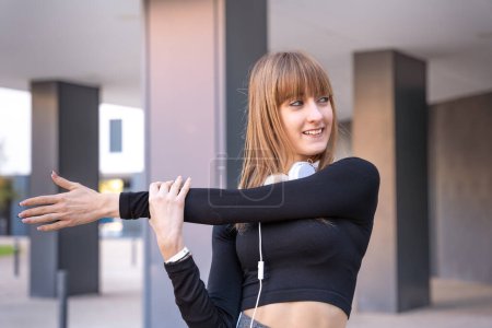 Téléchargez les photos : Jeune femme sportive portant des écouteurs de sport faisant des étirements souriant en regardant la caméra à l'extérieur. - en image libre de droit