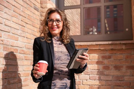 Photo for Portrait of smiling middle aged business woman with mobile phone and coffee in hands and glasses outdoors. - Royalty Free Image