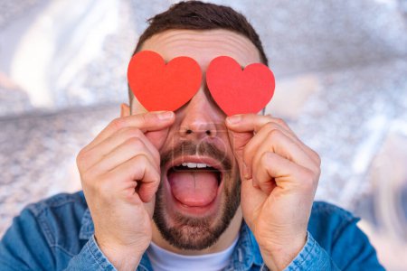 Foto de Hombre sonriente con humor sostiene dos corazones en los ojos en el Día de San Valentín. - Imagen libre de derechos