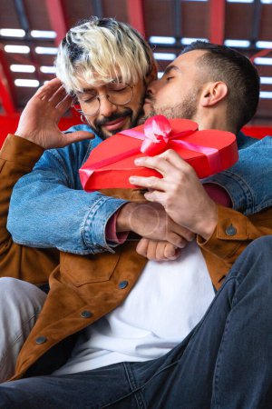 Foto de Hermosa pareja homosexual en casa disfrutando de tiempo juntos mientras celebran el Día de San Valentín con una caja de regalo en forma de corazón en la mano. besando con amor - Imagen libre de derechos
