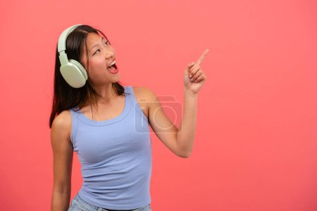Photo for Portrait of a beautiful young Asian woman wearing headphones pointing one hand with finger on the top left with smiling face. The girl standing in the studio. Advertising and presentation concept. - Royalty Free Image