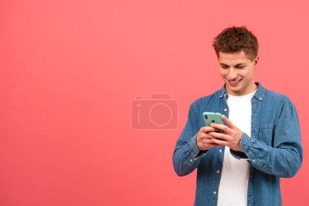 Téléchargez les photos : Portrait d'homme heureux regardant le téléphone mobile, souriant, textos sur fond rose. Concept d'émotions humaines, expression faciale. Espace de copie pour la publicité - en image libre de droit