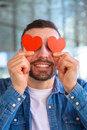 Photo for Smiling man with humor holds two hearts in eyes on Valentines Day. - Royalty Free Image