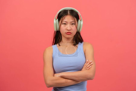 Téléchargez les photos : Femme chinoise en colère avec des écouteurs sur fond rose, avec les bras croisés regardant la caméra - en image libre de droit