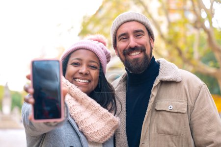 Téléchargez les photos : Couple touristique multiracial montre un smartphone avec un écran vide pour la publicité ou la présentation, regarder la caméra., sourire. Concept Gadget - en image libre de droit