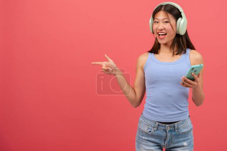 Foto de Mujer asiática joven con auriculares, señalando con el dedo el lado izquierdo de la foto con una cara sorprendida. La chica de pie con teléfono en el estudio. Concepto de publicidad y presentación. - Imagen libre de derechos
