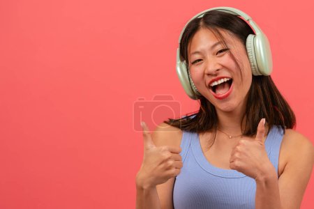Téléchargez les photos : Belle jeune femme asiatique portant des écouteurs pointant les doigts vers le haut sur le côté droit de la photo avec un visage heureux. La fille debout dans le studio. Concept de présentation publicitaire - en image libre de droit