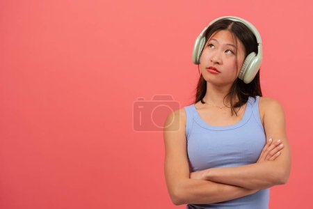 Téléchargez les photos : Femme chinoise en colère avec des écouteurs sur fond rose, les bras croisés regardant vers le haut - en image libre de droit