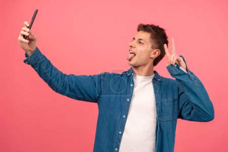 Photo for Portrait of a cheerful boy taking a selfie and making the peace sign on a pink background. Isolated. - Royalty Free Image