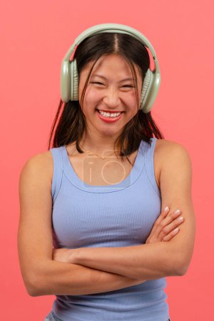 Téléchargez les photos : Heureuse femme chinoise avec écouteurs sur fond rose, les bras croisés en regardant la caméra - en image libre de droit