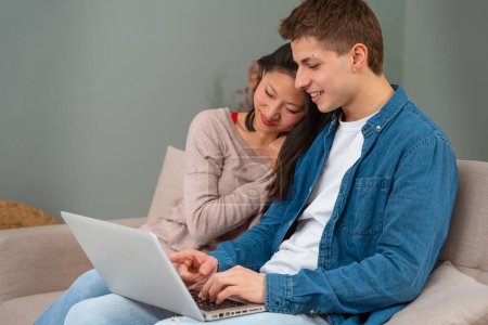 Photo for Smiling young multiracial couple sit embraced on sofa at home using laptop browsing wireless internet on device. A multiethnic man and woman have fun browsing the Internet on a laptop. - Royalty Free Image