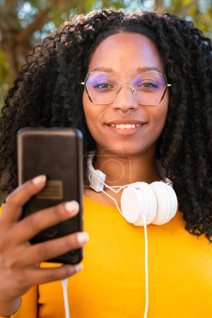Photo for Beautiful black woman with curly hair using her smartphone to chat with friend looking at camera connected concept. - Royalty Free Image