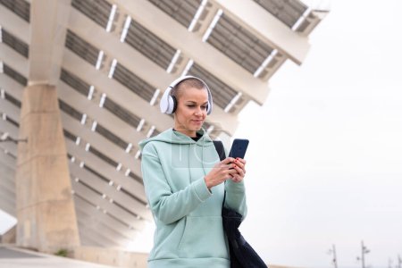 An athletic woman with headphones tired from exercising takes a break to use a cell phone messaging app.
