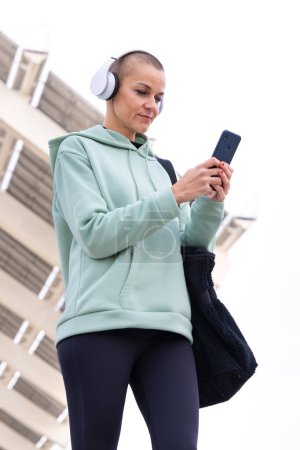 Photo for An athletic woman with headphones tired from exercising takes a break to use a cell phone messaging app. - Royalty Free Image