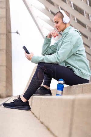 Téléchargez les photos : Une femme sportive avec un casque fatigué de l'exercice s'assoit pour prendre une pause aquatique tout en utilisant une application de téléphone portable. - en image libre de droit
