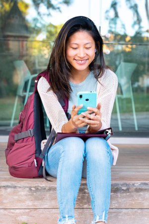 Foto de Hermosa estudiante adolescente china sonriendo usando una aplicación de redes sociales en un teléfono inteligente al aire libre. Chica charlando en el teléfono móvil - Imagen libre de derechos