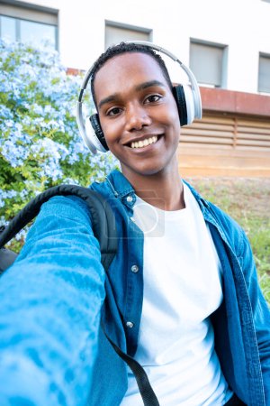 Foto de Joven estudiante negro de pie al aire libre en la universidad con auriculares que hacen un autorretrato con el teléfono inteligente.Concepto de vida universitaria - Imagen libre de derechos