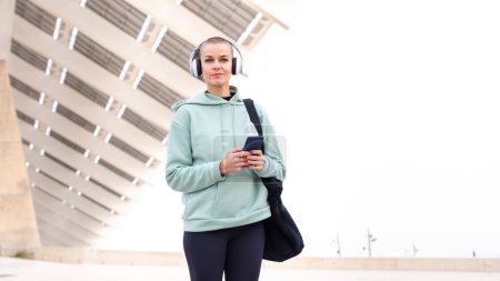 An athletic woman with headphones tired from exercising takes a break to use a cell phone messaging app looking at camera.