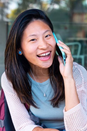 Foto de Hermosa estudiante adolescente china sonriendo, teniendo una conversación en un teléfono móvil al aire libre. Chica sentada charlando en el teléfono celular - Imagen libre de derechos