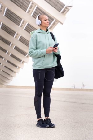 An athletic woman with headphones tired from exercising takes a break to use a cell phone messaging app outdoors.