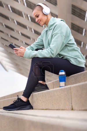 Téléchargez les photos : Une femme sportive avec un casque fatigué de l'exercice s'assoit pour prendre une pause aquatique tout en utilisant une application de téléphone portable. - en image libre de droit