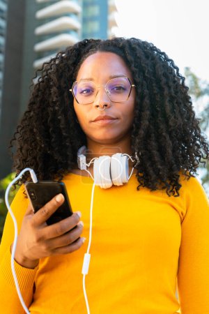 Photo for Beautiful serious black woman with curly hair using her smartphone to chat with friend looking at camera connected concept. - Royalty Free Image