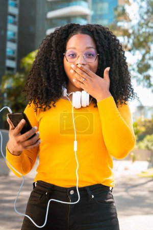 Téléchargez les photos : Belle femme noire surprise avec des cheveux bouclés à l'aide de son smartphone pour discuter avec un ami en regardant la caméra concept connecté. - en image libre de droit