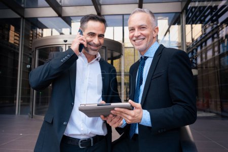 Photo for Two middle aged adult men in suits have a business call and the other one is holding digital tablet looking at camera outdoors. - Royalty Free Image
