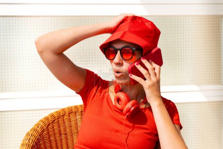 Foto de Elegante mujer joven caucásica sonriendo haciendo una llamada con un teléfono celular con gafas y sombrero rojo Una hermosa mujer caucásica con sombrero rojo y gafas sonriendo al aire libre. - Imagen libre de derechos
