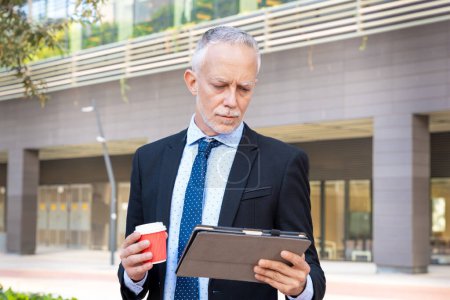 Photo for Satisfied with the results, the businessman walks down the street outside the office building, a mature boss holds adigital tablet and coffee in his hands, writes messages and reads news online, - Royalty Free Image