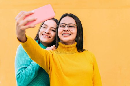 Téléchargez les photos : Deux jolies jeunes femmes hispaniques souriantes en pull bleu et jaune avec des bretelles sur les dents prenant un selfie. concept d'amitié. - en image libre de droit
