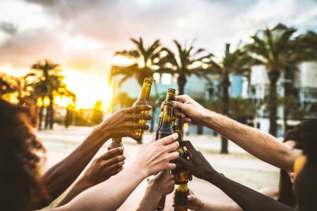 Téléchargez les photos : Groupe d'amis multiethniques heureux faisant la fête à l'extérieur pour célébrer les bouteilles de bière grillées au coucher du soleil. Concept de célébration et de bonheur - en image libre de droit