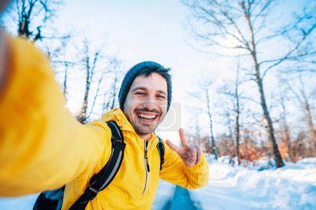 Foto de Hombre feliz con mochila tomando retrato de selfie en bosque helado de invierno - Smiling hiker climbing mountains - Concepto de deporte, viajes y tecnología - Imagen libre de derechos