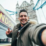 Smiling man taking selfie portrait during travel in London, England - Young tourist male taking memory pic with iconic england landmark - Happy people wandering around Europe concept