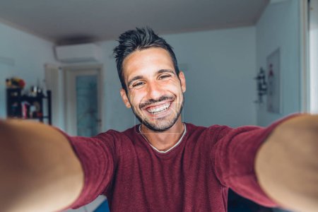 Foto de Feliz joven guapo hombre milenario tomando una selfie sonriendo a la cámara en la sala de estar en casa - Imagen libre de derechos
