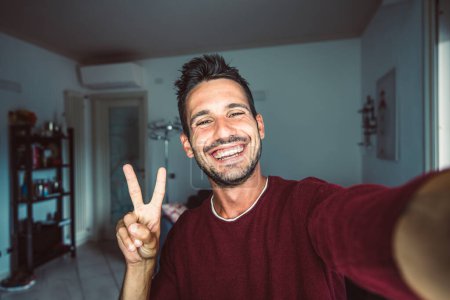Foto de Feliz joven guapo hombre milenario tomando una selfie sonriendo a la cámara en la sala de estar en casa - Imagen libre de derechos