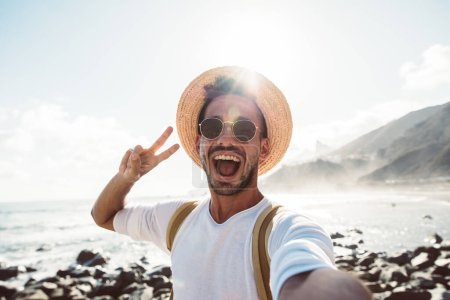 Téléchargez les photos : Jeune homme avec sac à dos prendre selfie à l'extérieur - Happy guy profiter des vacances d'été à la plage - Millénaire montrant symbole des mains de victoire à l'appareil photo - en image libre de droit