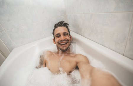 Photo for Happy handsome man take a selfie inside bathtub in the bathroom of the hotel on holiday - travel freedom beauty concept - Royalty Free Image