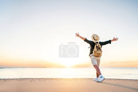 Photo for Happy man wearing hat and backpack raising arms up on the beach at sunset - Delightful man enjoying peaceful moment walking outdoors - Wellness, healthcare, traveling and mental health concept - Royalty Free Image